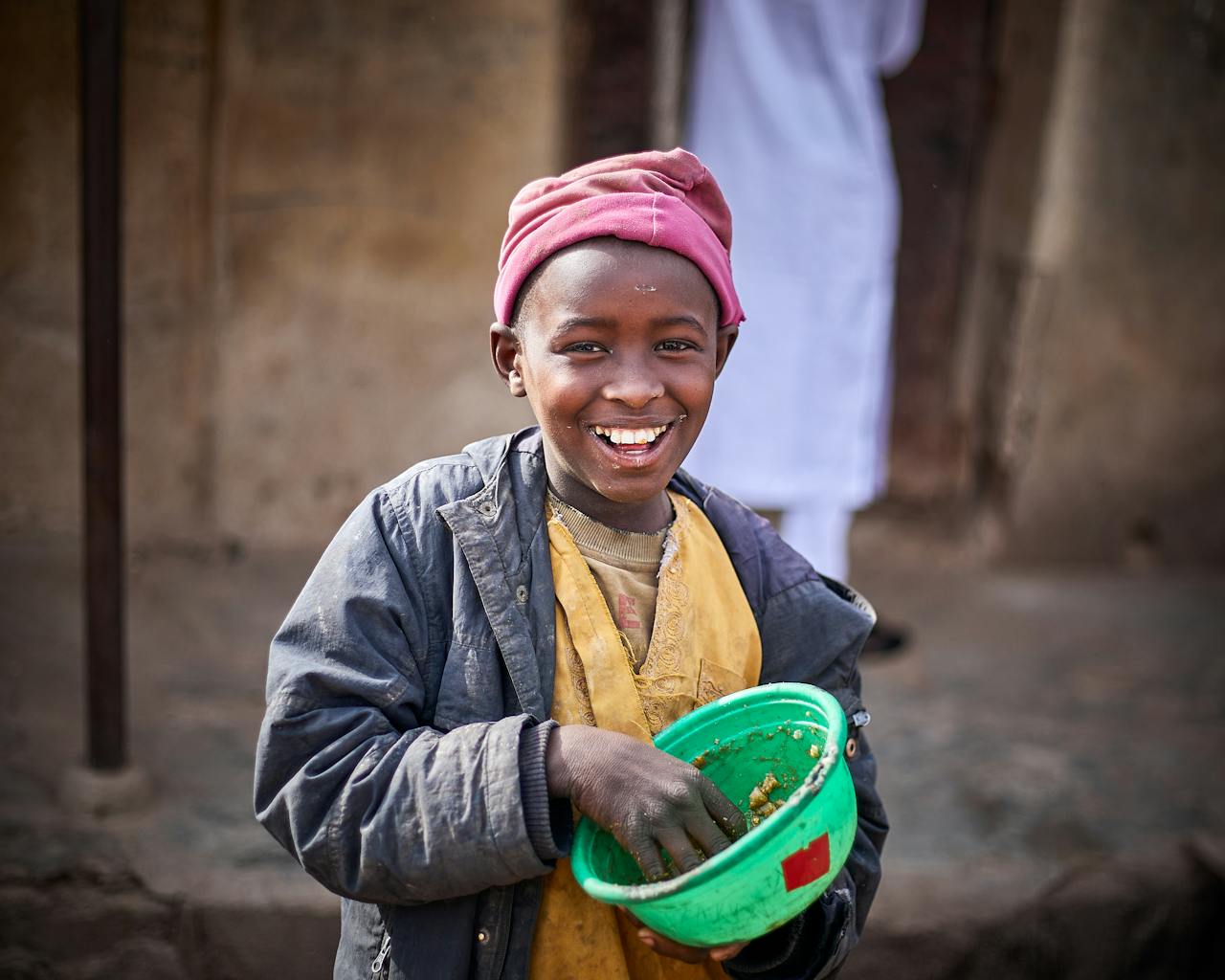 about-01 Happy Nigerian child holding a bowl outdoors in Kaduna, wearing a colorful beret and jacket.