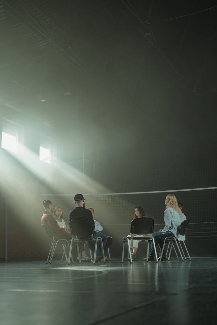 hero-about A group of adults in a therapy session, sitting in a dimly lit room with sun rays streaming in.